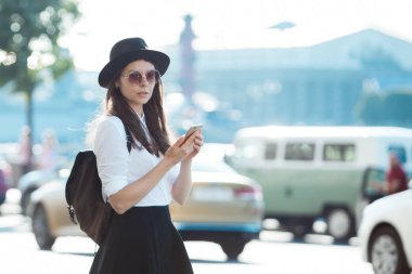 bir şapkalı genç kadın ve bir sırt çantası ile şehirde yürür ve bir akıllı telefon kullanır.