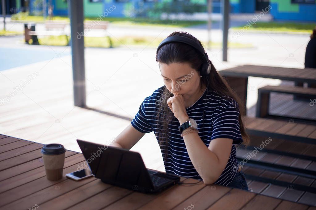 Pensive girl student, brunette, in headphones with a smartphone, laptop and paper cup of coffee sits at a wooden table.