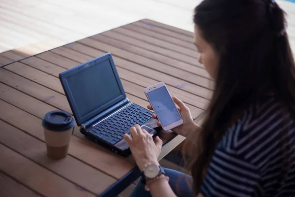 Брюнетка с большими наручными часами, полуразвернутая, работает за ноутбуком, стоящим на столе. Рядом находится смартфон. . — стоковое фото