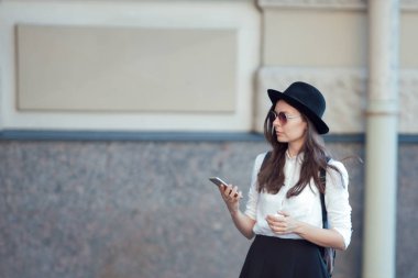 bir şapkalı genç kadın ve bir sırt çantası ile şehirde yürür ve bir akıllı telefon kullanır.