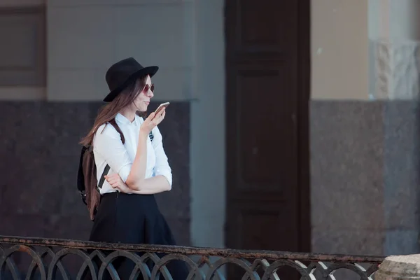 Mujer joven con un sombrero y una camisa blanca camina por la ciudad y utiliza un teléfono inteligente . —  Fotos de Stock