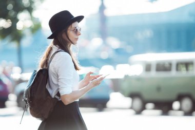 bir şapkalı genç kadın ve bir sırt çantası ile şehirde yürür ve bir akıllı telefon kullanır.