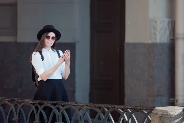 Giovane donna con un cappello e una camicia bianca cammina in città e utilizza uno smartphone . — Foto Stock
