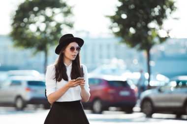 bir şapkalı genç kadın ve bir sırt çantası ile şehirde yürür ve bir akıllı telefon kullanır.