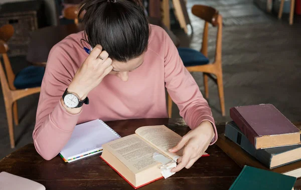 Exam preparation. Student girl in glasses and a pink sweater leaned over books, engaged in an old library at the table.