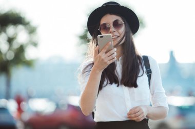 bir şapkalı genç kadın ve bir sırt çantası ile şehirde yürür ve bir akıllı telefon kullanır.