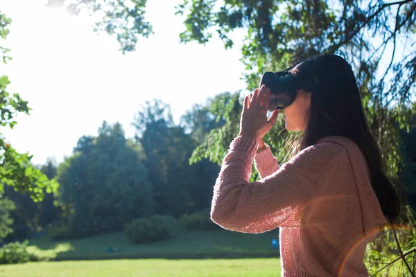 Virtual forest. A young woman in VR glasses stands under the trees in a clearing, on sunny day, touches the air.