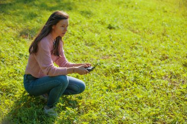 Parkta bir açıklıkta çömelmiş bir kadın kontrol paneli monitöründen uçan bir insansız hava aracını izliyor..
