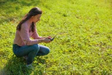 Parkta bir açıklıkta çömelmiş bir kadın kontrol paneli monitöründen uçan bir insansız hava aracını izliyor..