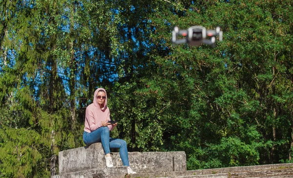 Una joven con capucha rosa y gafas de sol sentada en un banco de piedra y operada por un avión teledirigido . —  Fotos de Stock