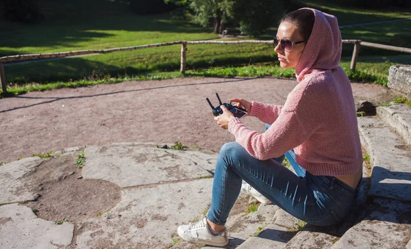 Молодая женщина в розовой толстовке и солнечных очках, сидящая на каменной скамейке и управляемая беспилотником с дистанционным управлением . — стоковое фото