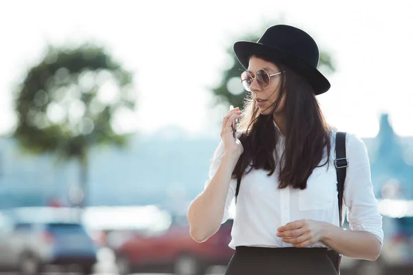 Jovem mulher em um chapéu e com uma mochila caminha na cidade e usa um smartphone . — Fotografia de Stock