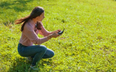 Parkta bir açıklıkta çömelmiş bir kadın kontrol paneli monitöründen uçan bir insansız hava aracını izliyor..