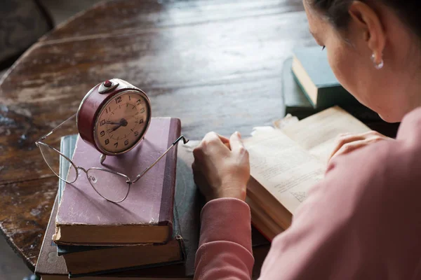 Demasiado poco tiempo. Reloj despertador parado en una pila de libros, junto a las gafas, frente a una chica inclinada sobre un libro . — Foto de Stock