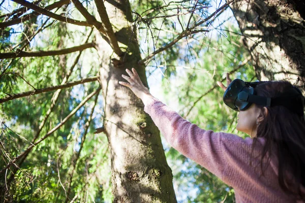 Virtual forest. A young woman among the trees, wearing virtual reality glasses, touches the boles and branches.