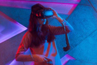 Sanal gerçeklik gözlük genç bir kadın neon süslemeleri ile duvar arka plan karşı, denetleyicileri kullanır.