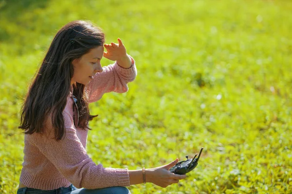 A woman squatting in a clearing in a park is watching a drone flying through a control panel monitor.