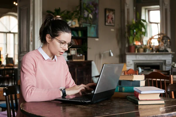 Exam preparation. Attractive brunette student girl in glasses and a pink sweater, with a slight smile sits at a laptop.