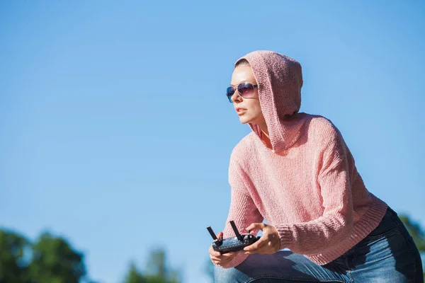Giovane donna che indossa occhiali da sole e cappuccio sulla testa tenendo un telecomando, guarda attentamente in lontananza contro . — Foto Stock