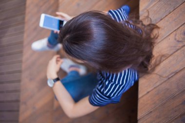 Büyük bir bilek saati ile esmer kız, ahşap adımlarla oturur ve bir akıllı telefon kontrol eder. Görünüm yukarıda açıkça.