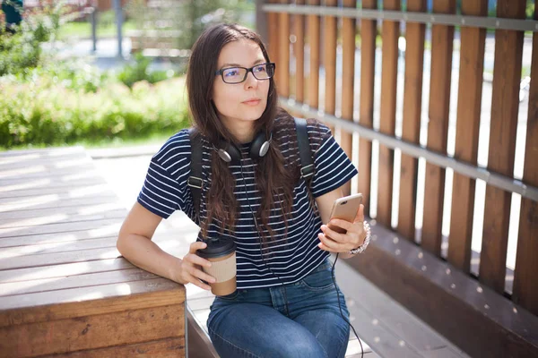 Hübsches Mädchen mit Brille, sitzt auf Bank, mit Tasse Kaffee und Smartphone und blickt nachdenklich in die Ferne. — Stockfoto
