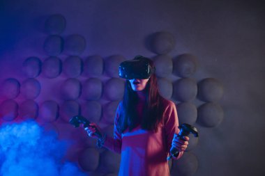 Birinci şahıs nişancı. Gözlüklü ve sanal gerçeklik kaskı olan bir kadın, silah gibi iki elinde de oyun kumandalarını kullanır..