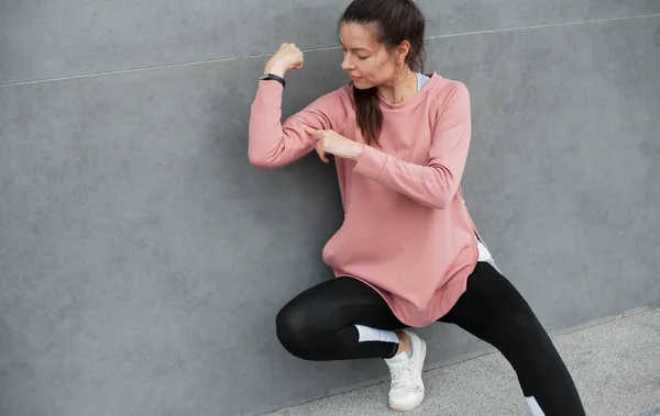 Cheerful, funny, fitness young woman resting after exercise, checking biceps, sitting against the wall.