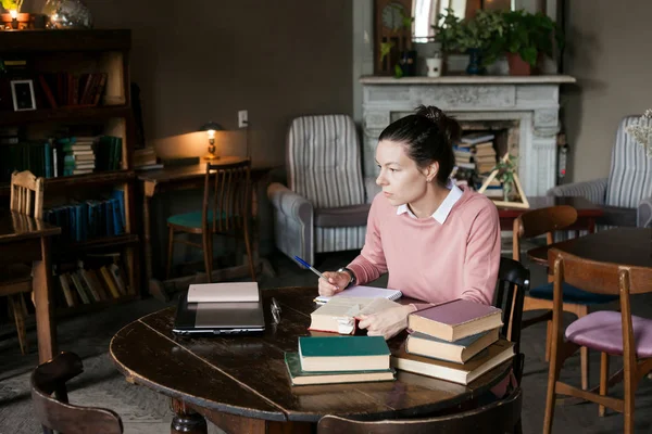 Exam preparation. Student girl in glasses and a pink sweater leaned over books, engaged in an old library at the table.