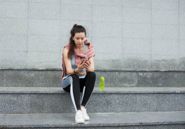 Spor üniforması atletik genç kadın oturur, bir ısınma sonrası akıllı telefon ve kablosuz kulaklık ile merdivenlerde dinlenir.