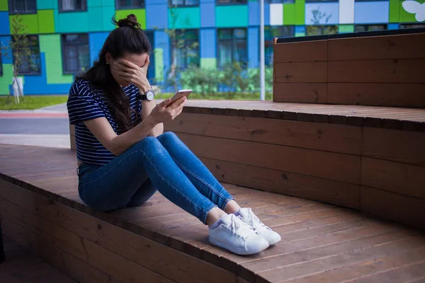 Молода дівчина з довгим волоссям, з телефоном, плаче, прикриває обличчя рукою, сидить з ногами на дерев'яних сходах . — стокове фото