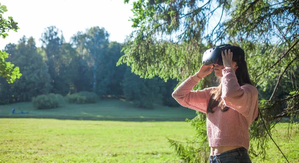 Virtual forest. A young woman in VR glasses stands under the trees in a clearing, on sunny day, touches the air.