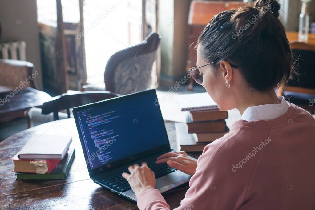 Hacking system. Excited girl with dark hair and in pink sweater and glasses writes code on a laptop in a library.