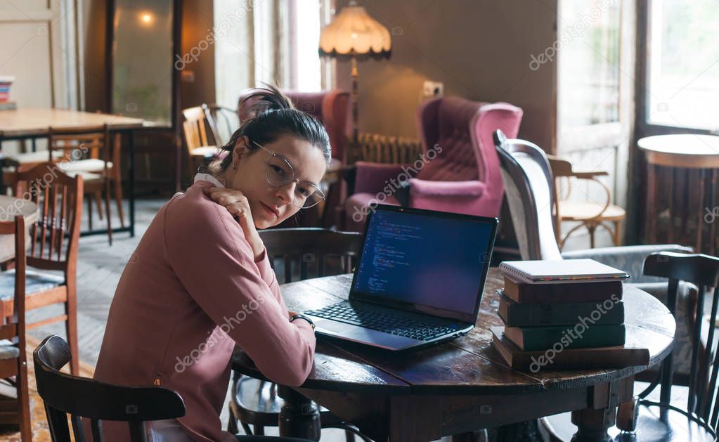 Site development. Smart girl with a soft smile, sits at desk in the library, in front of her is laptop with a site code.