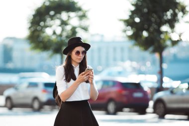 şapkalı ve beyaz gömlekli genç kadın şehirde yürür ve akıllı telefon kullanır.