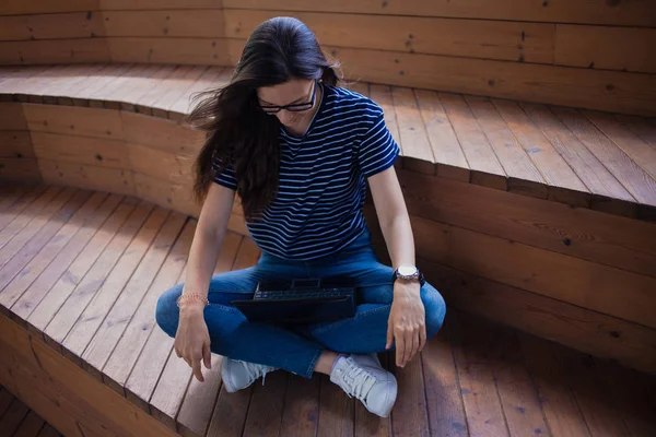 O fată brunetă, un student cu păr lung, cu ochelari așezați cu picioarele încrucișate pe trepte de lemn și tastând pe laptop . — Fotografie, imagine de stoc