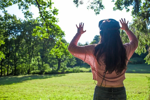 Virtual forest. Young woman in VR glasses stands under the trees in a clearing, on sunny summer day and touches the air.
