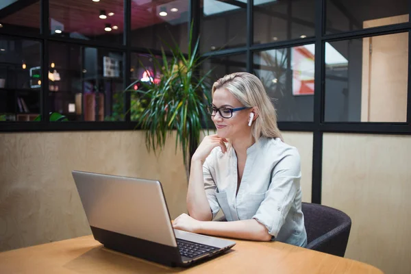 Jonge blonde vrouw zit op haar laptop en maakt gebruik van draadloze hoofdtelefoon. — Stockfoto