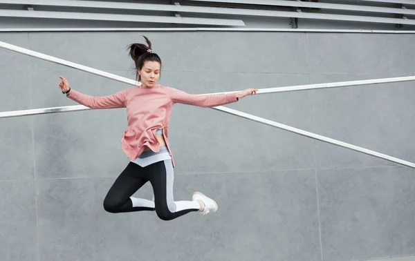 Een atletische jonge vrouw springt, doet acrobatiek, ballet, is actief betrokken bij sport, tegen een betonnen muur. — Stockfoto