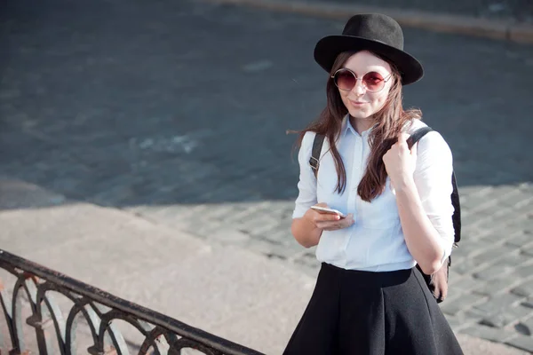 Jonge vrouw in een hoed en een wit shirt wandelingen in de stad en maakt gebruik van een smartphone. — Stockfoto