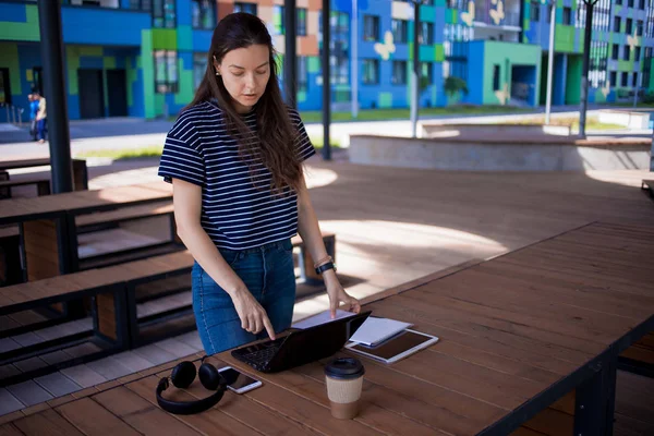 Серьезная девушка стоит перед ноутбуком, внимательно изучая документы и бумаги, которые она держит в руках. . — стоковое фото