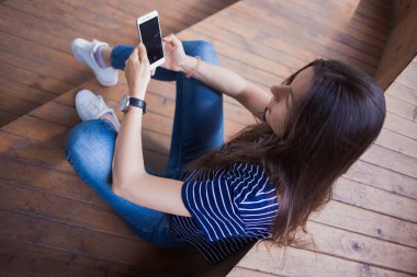 Büyük kol saati olan esmer bir kız, ahşap merdivenlerde oturur ve akıllı telefonu kontrol eder. Manzara yukarıdan..