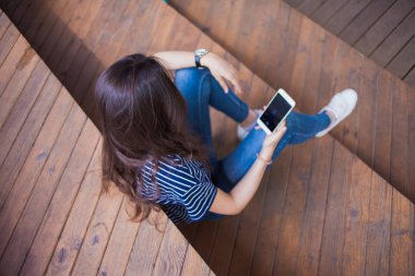 Büyük bir bilek saati ile esmer kız, ahşap adımlarla oturur ve bir akıllı telefon kontrol eder. Görünüm yukarıda açıkça.