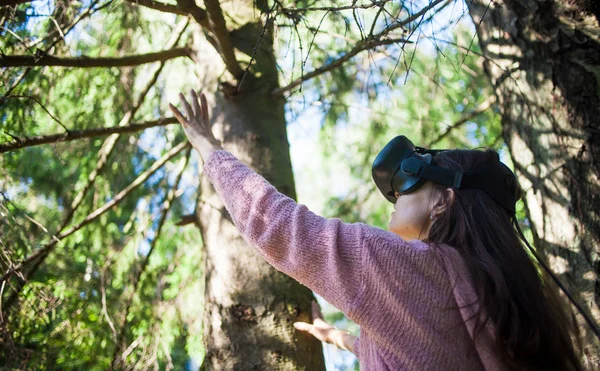 Virtual forest. A young woman among the trees, wearing virtual reality glasses, touches the boles and branches.