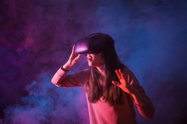 Genç bir kadın, arka planda dumanlı karanlık bir arka plan için gözlük ve sanal gerçeklik kaskı kullanıyor. Gözlükleri düzeltir.