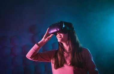 Genç bir kadın, arka planda dumanlı karanlık bir arka plan için gözlük ve sanal gerçeklik kaskı kullanıyor. Gözlükleri düzeltir.