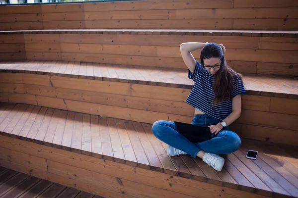 Una seria studentessa bruna, si siede su gradini di legno, con un computer portatile aperto, digitando su una tastiera, impara online . — Foto Stock