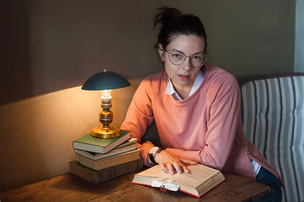 Een ijverige student. Meisje in bril met een dik boek met veel bladwijzers, aan een kleine tafel, onder een warme lamp. — Stockfoto