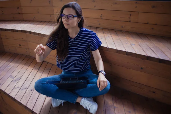 O fată brunetă, un student cu păr lung, cu ochelari așezați cu picioarele încrucișate pe trepte de lemn și tastând pe laptop . — Fotografie, imagine de stoc