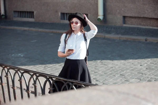 Mujer joven con un sombrero y una camisa blanca camina por la ciudad y utiliza un teléfono inteligente . — Foto de Stock