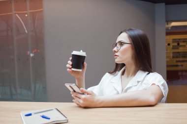 Öğrenci kız masada oturuyor ve akıllı telefonunda bir uygulama kullanıyor. Genç bir esmer, gözlüklü ve beyaz gömlekli.